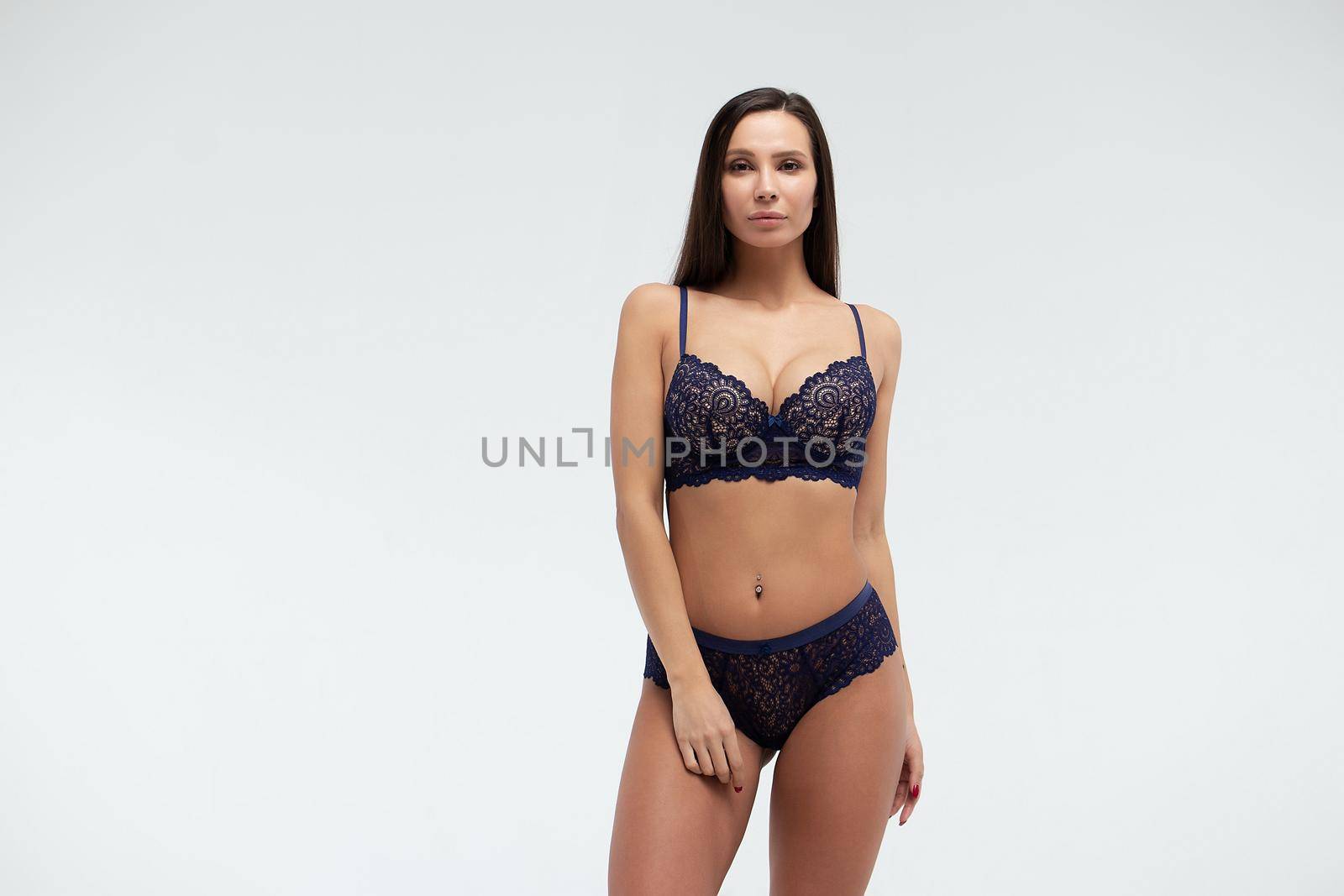 Serene slim female in dark blue lace underwear standing on white background in studio and looking away