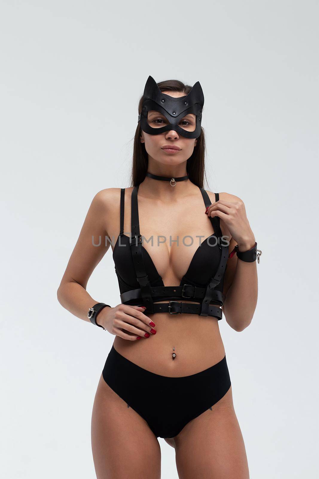 Slim seductive female wearing black mask and sexy underwear standing with hands on waist on white background and looking at camera