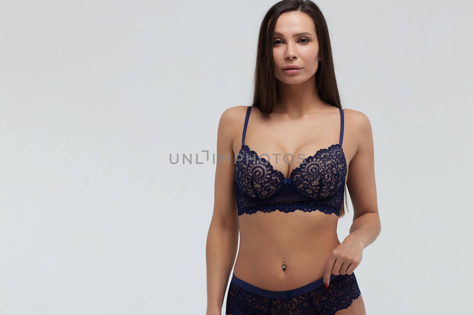 Serene slim female in dark blue lace underwear standing on white background in studio and looking away