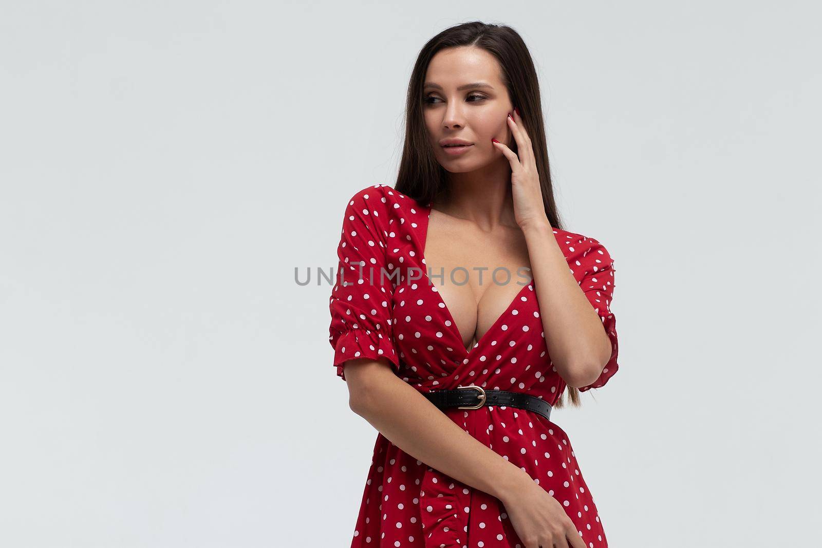 Sexy female wearing red short polka dot dress standing on white background in studio and touching hair while looking at camera