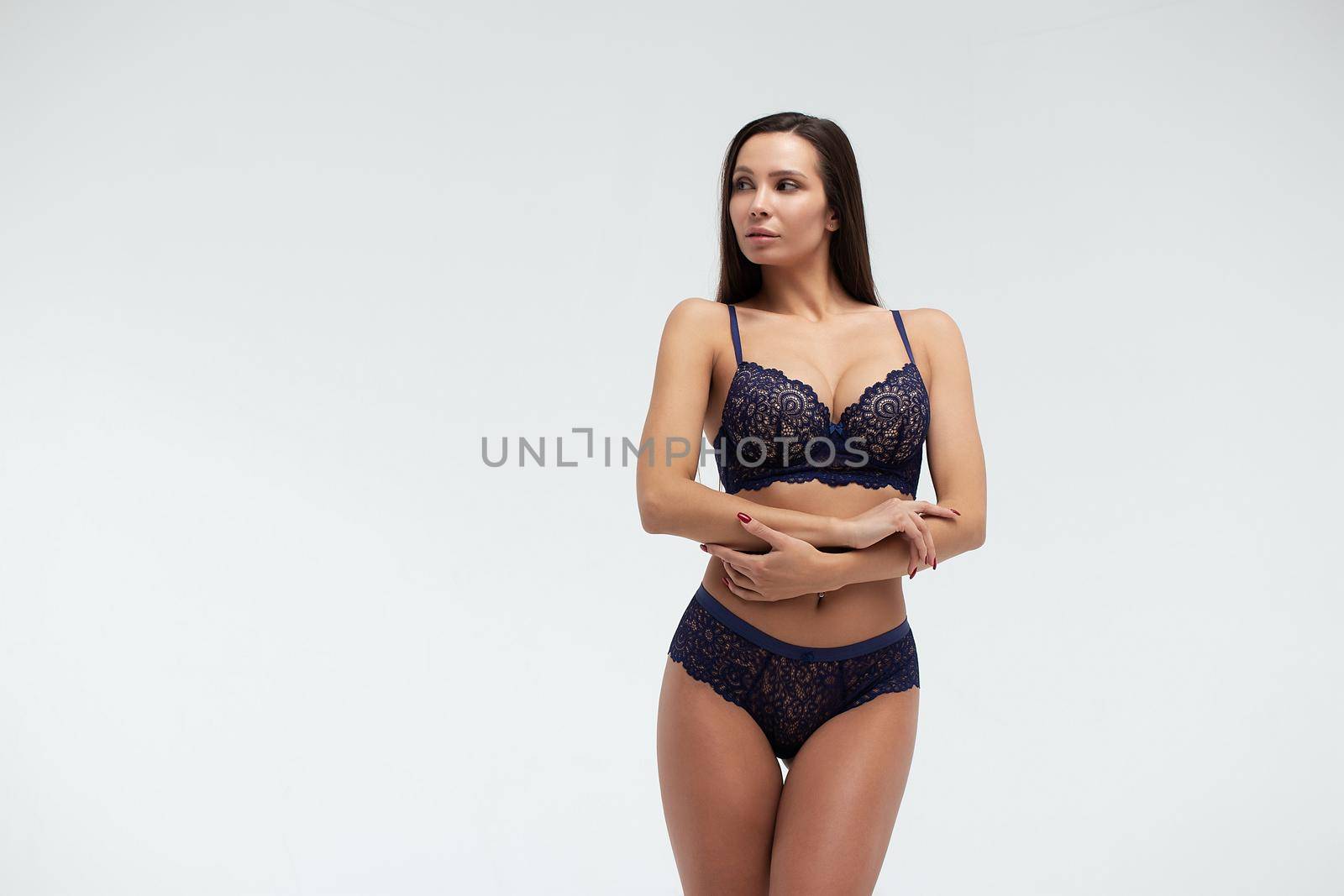 Serene slim female in dark blue lace underwear standing on white background in studio and looking away