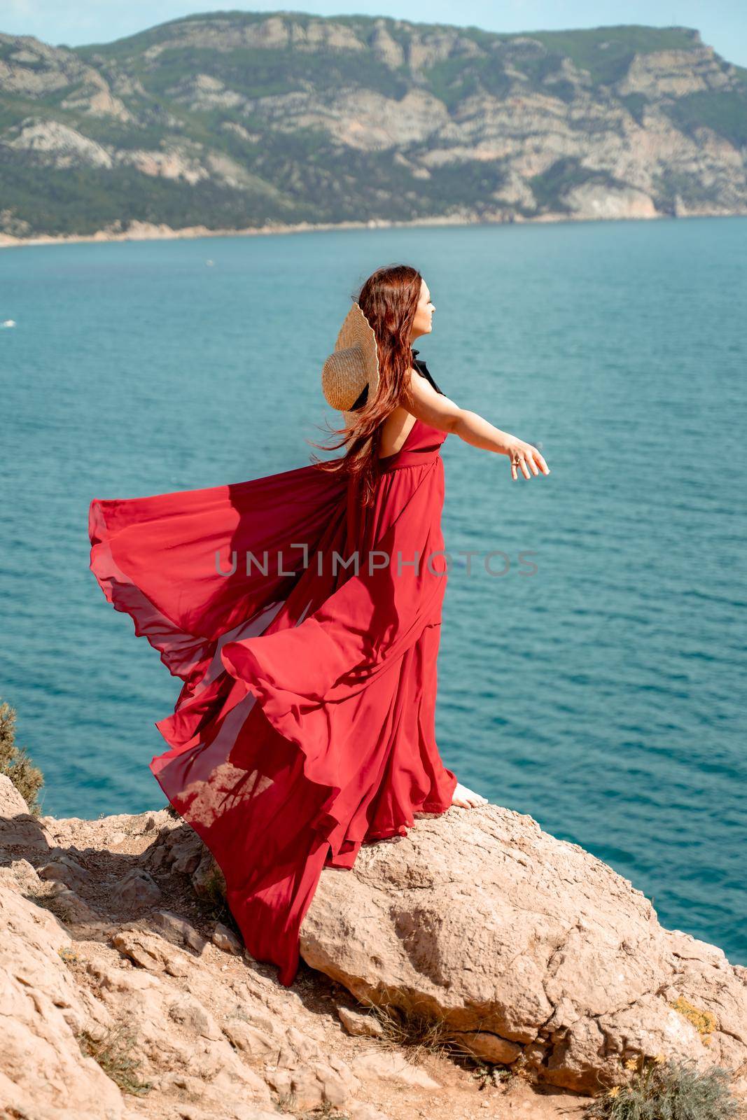A woman, a side view in a red flying dress fluttering in the wind, a girl in a fluttering dress on the background of the sea. A straw hat hangs at the back of the neck. The concept of a vacation at sea, a photo shoot by the sea, relaxation