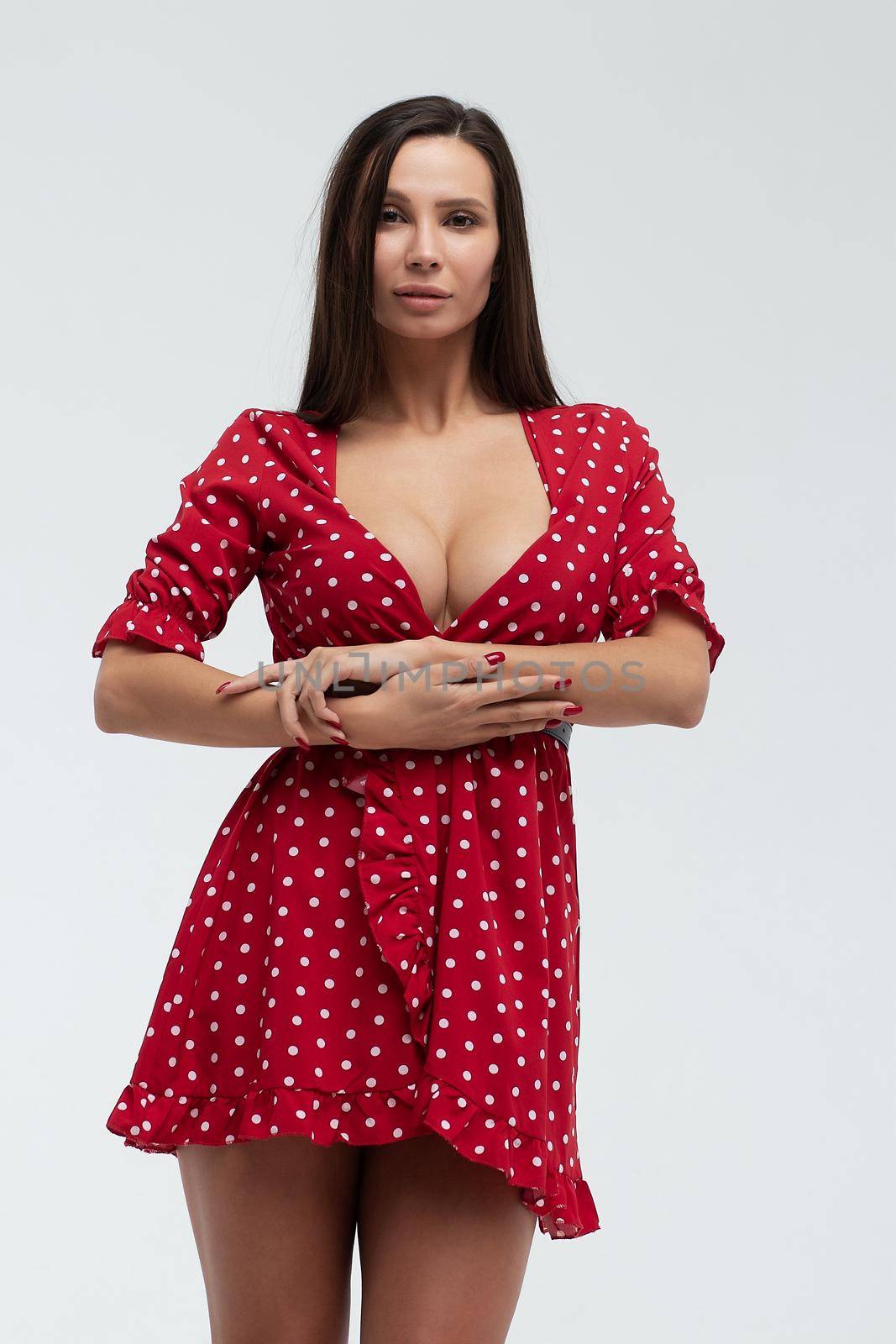 Sexy female wearing red short polka dot dress standing on white background in studio and touching hair while looking at camera