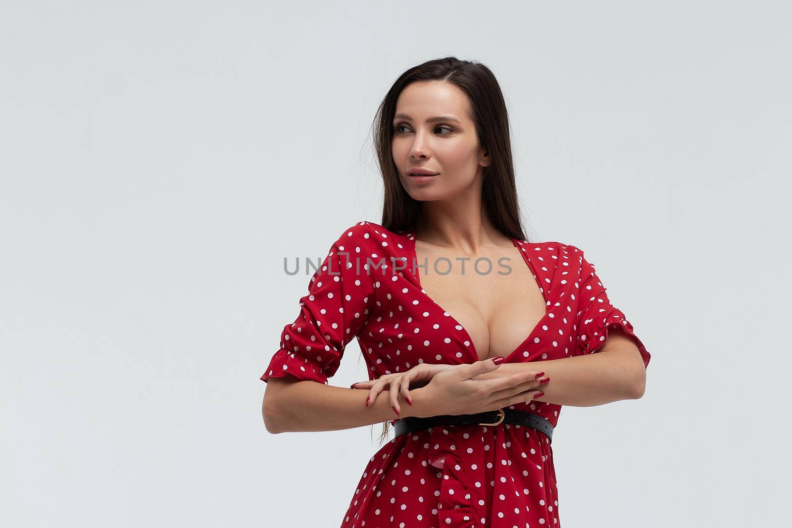 Sexy female wearing red short polka dot dress standing on white background in studio and touching hair while looking at camera