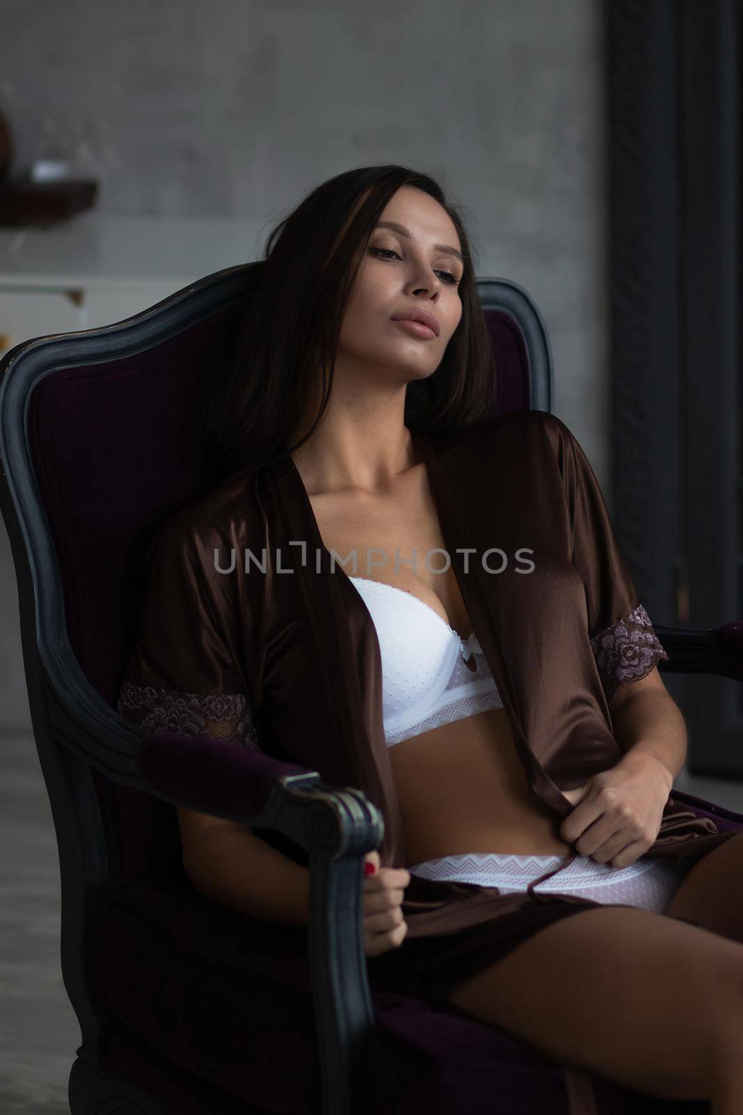Sensual young female with long dark hair in stylish dress with decollete relaxing in comfortable armchair and looking away