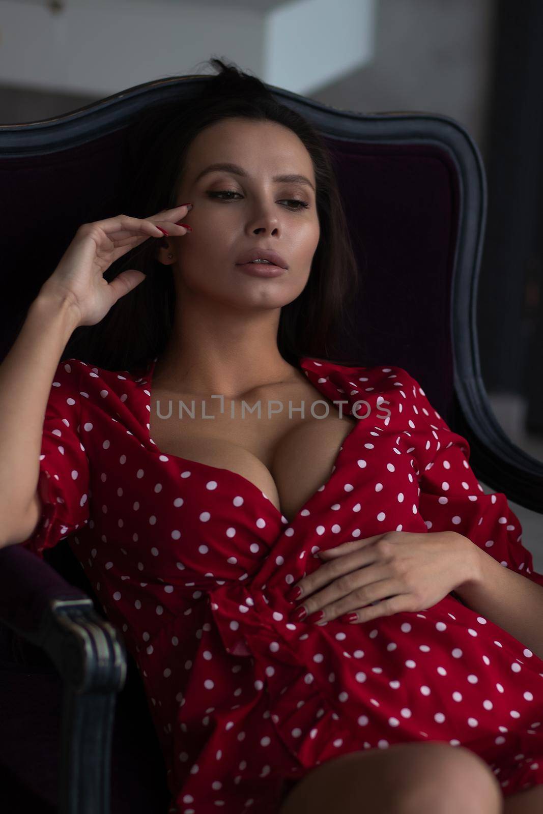 Sensual young female with long dark hair in stylish dress with decollete relaxing in comfortable armchair and looking away