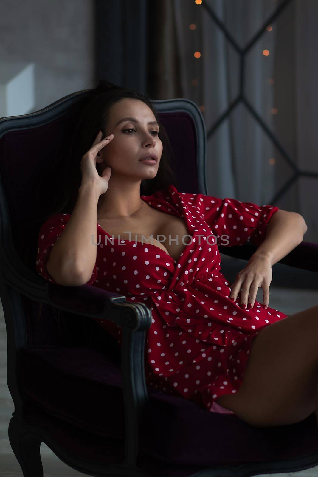 Sensual young female with long dark hair in stylish dress with decollete relaxing in comfortable armchair and looking away