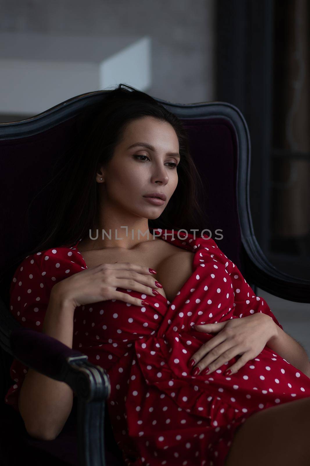 Sensual young female with long dark hair in stylish dress with decollete relaxing in comfortable armchair and looking away