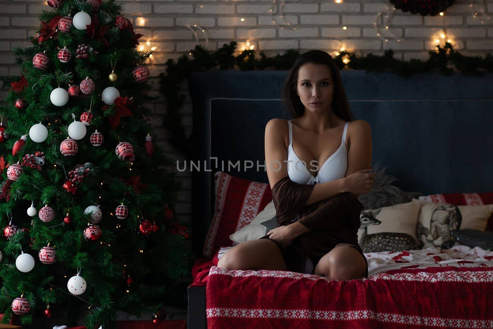 Self assured young lady in brown silk robe and white bra sitting on comfortable bed in cozy room decorated with glowing garlands and wreath during Christmas holidays