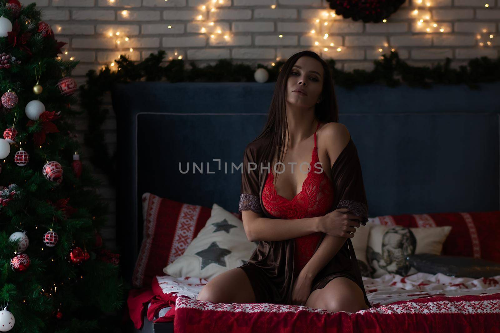 Self assured young lady in brown silk robe and red bodysuit sitting on comfortable bed in cozy room decorated with glowing garlands and wreath during Christmas holidays.