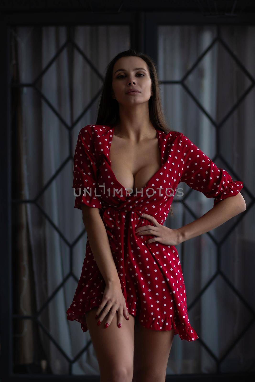 Self esteem young female with long hair in stylish mini dress with decollete standing in dark bedroom with hand on waist and looking at camera