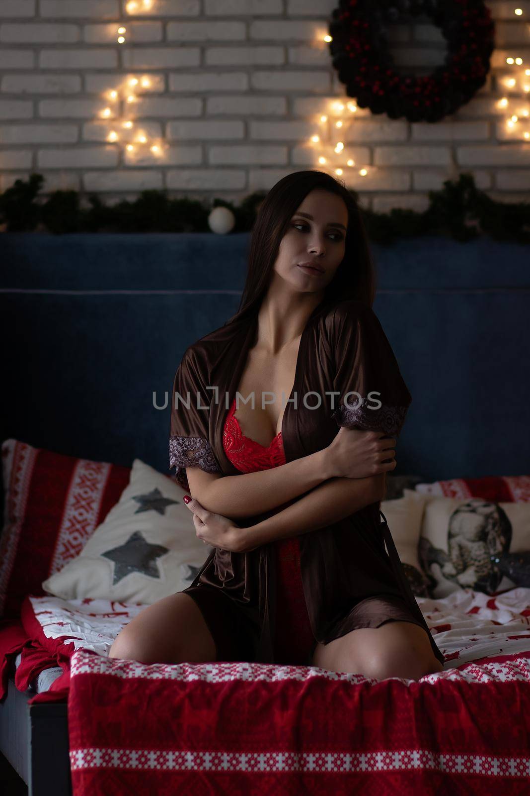 Self assured young lady in brown silk robe and white bra sitting on comfortable bed in cozy room decorated with glowing garlands and wreath during Christmas holidays