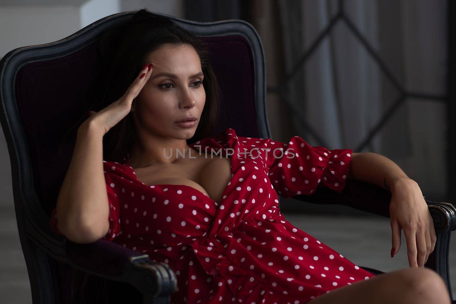 Sensual young female with long dark hair in stylish dress with decollete relaxing in comfortable armchair and looking away