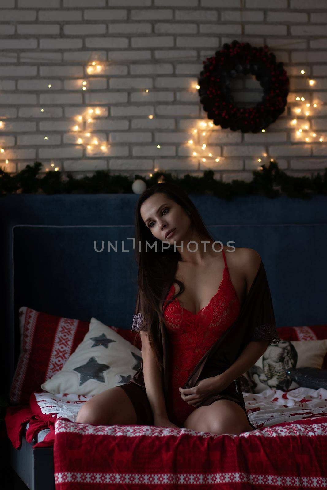 Self assured young lady in brown silk robe and white bra sitting on comfortable bed in cozy room decorated with glowing garlands and wreath during Christmas holidays