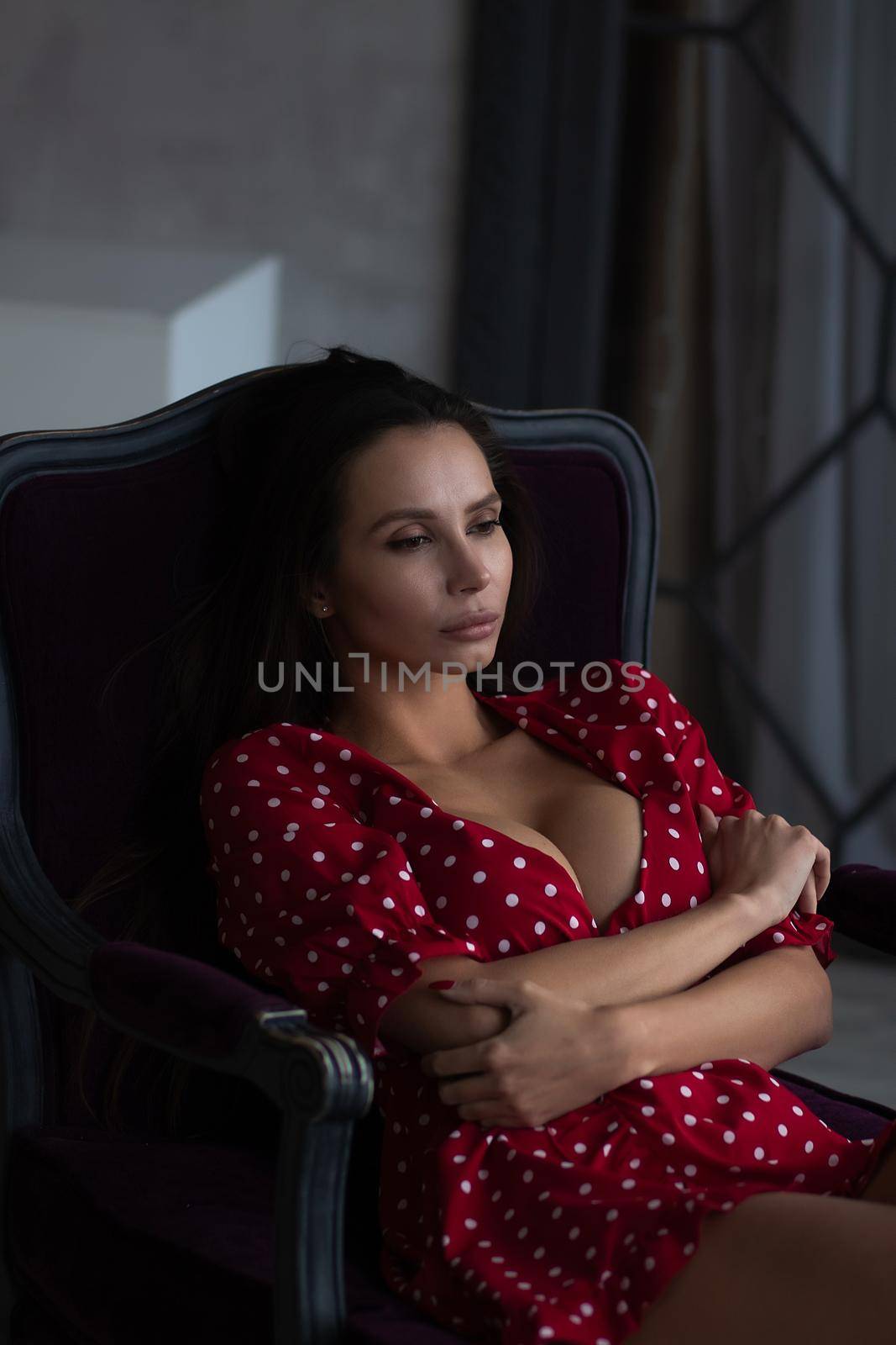 Sensual young female with long dark hair in stylish dress with decollete relaxing in comfortable armchair and looking away