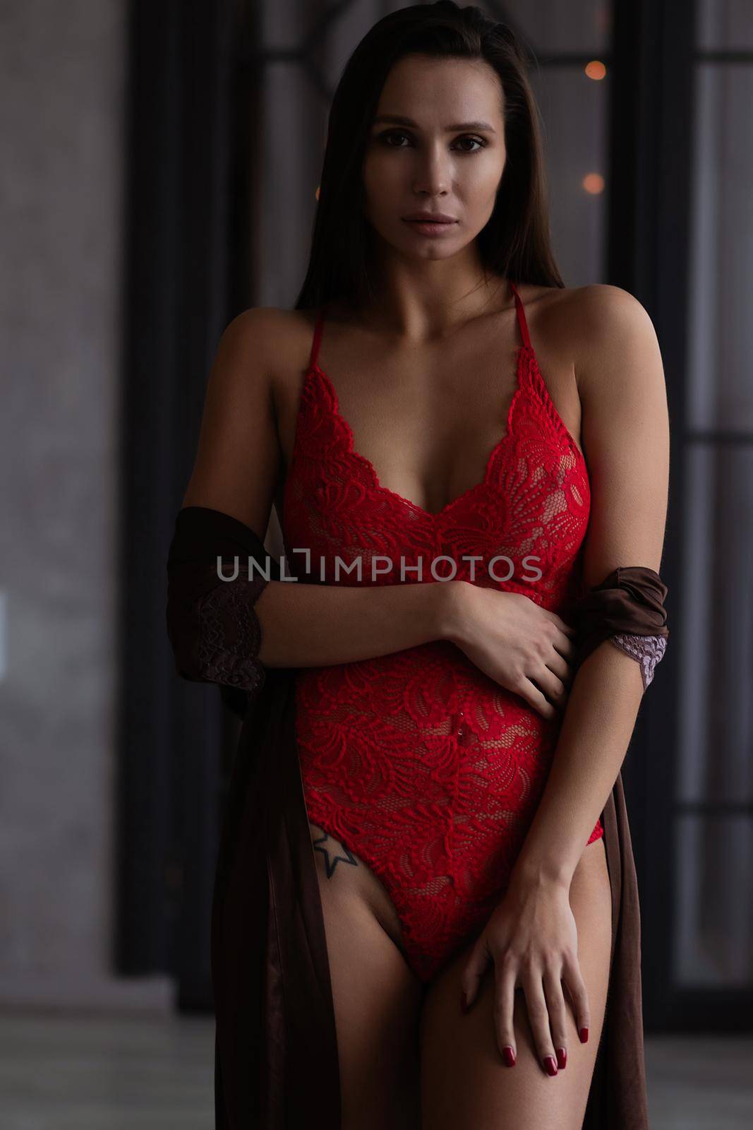 Sensual curvy female in red lingerie and brown silk robe standing gracefully in dark room and looking at camera