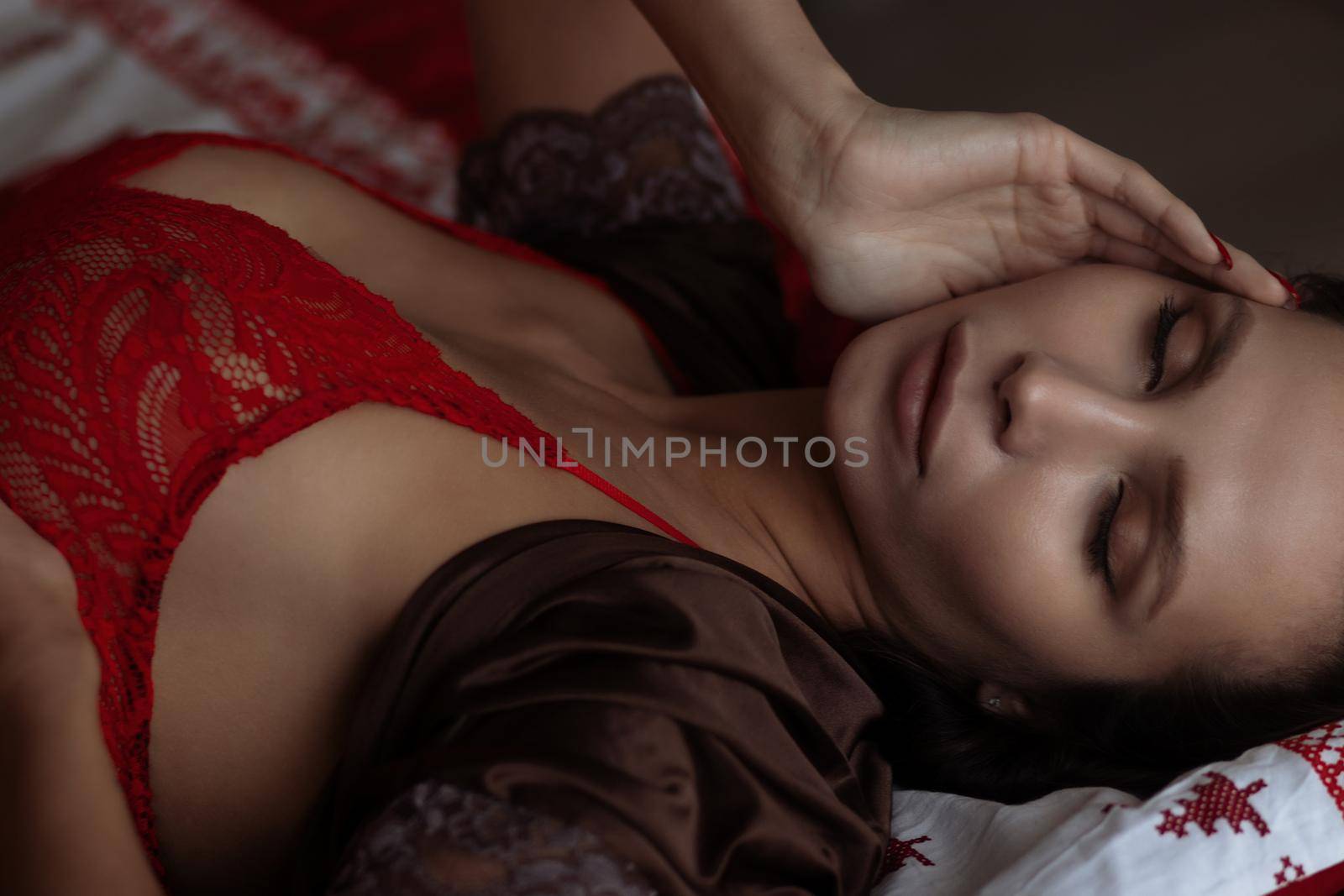 Side view of sexual young female with long dark hair in elegant red bra lying on comfortable bed with closed eyes near decorated Christmas tree