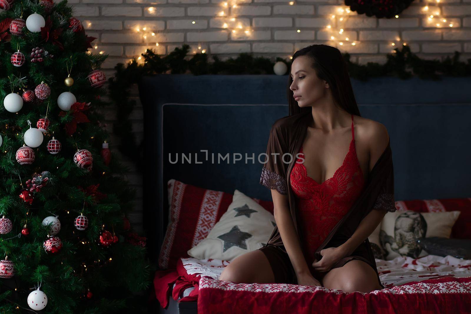 Self assured young lady in brown silk robe and red bodysuit sitting on comfortable bed in cozy room decorated with glowing garlands and wreath during Christmas holidays