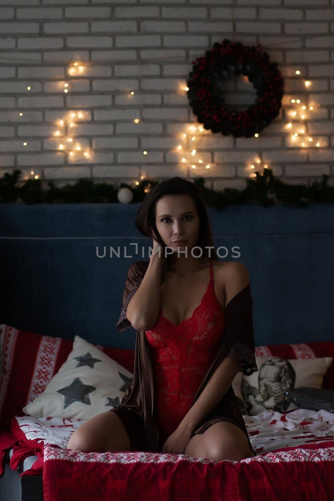 Self assured young lady in brown silk robe and red bodysuit sitting on comfortable bed in cozy room decorated with glowing garlands and wreath during Christmas holidays