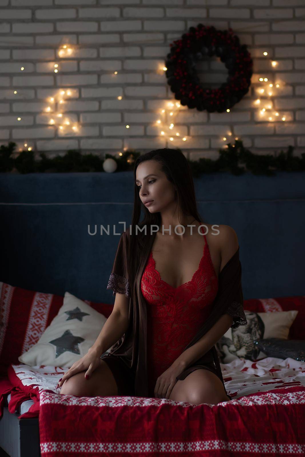 Self assured young lady in brown silk robe and red bodysuit sitting on comfortable bed in cozy room decorated with glowing garlands and wreath during Christmas holidays