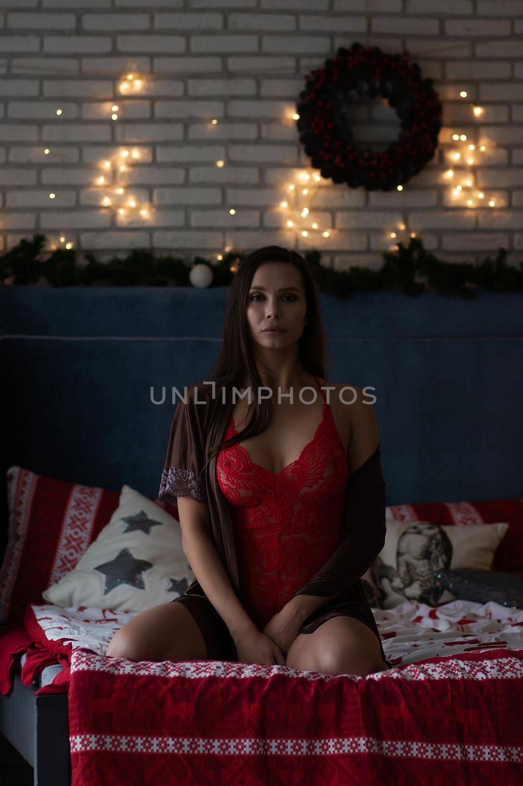 Self assured young lady in brown silk robe and red bodysuit sitting on comfortable bed in cozy room decorated with glowing garlands and wreath during Christmas holidays