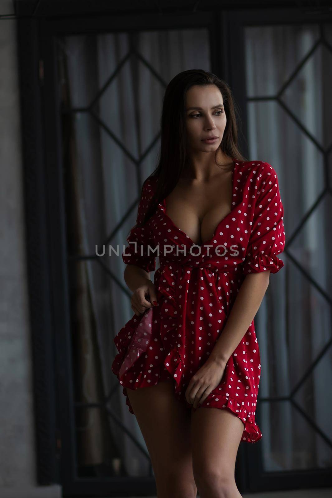 Self esteem young female with long hair in stylish mini dress with decollete standing in dark bedroom with hand on waist and looking at camera