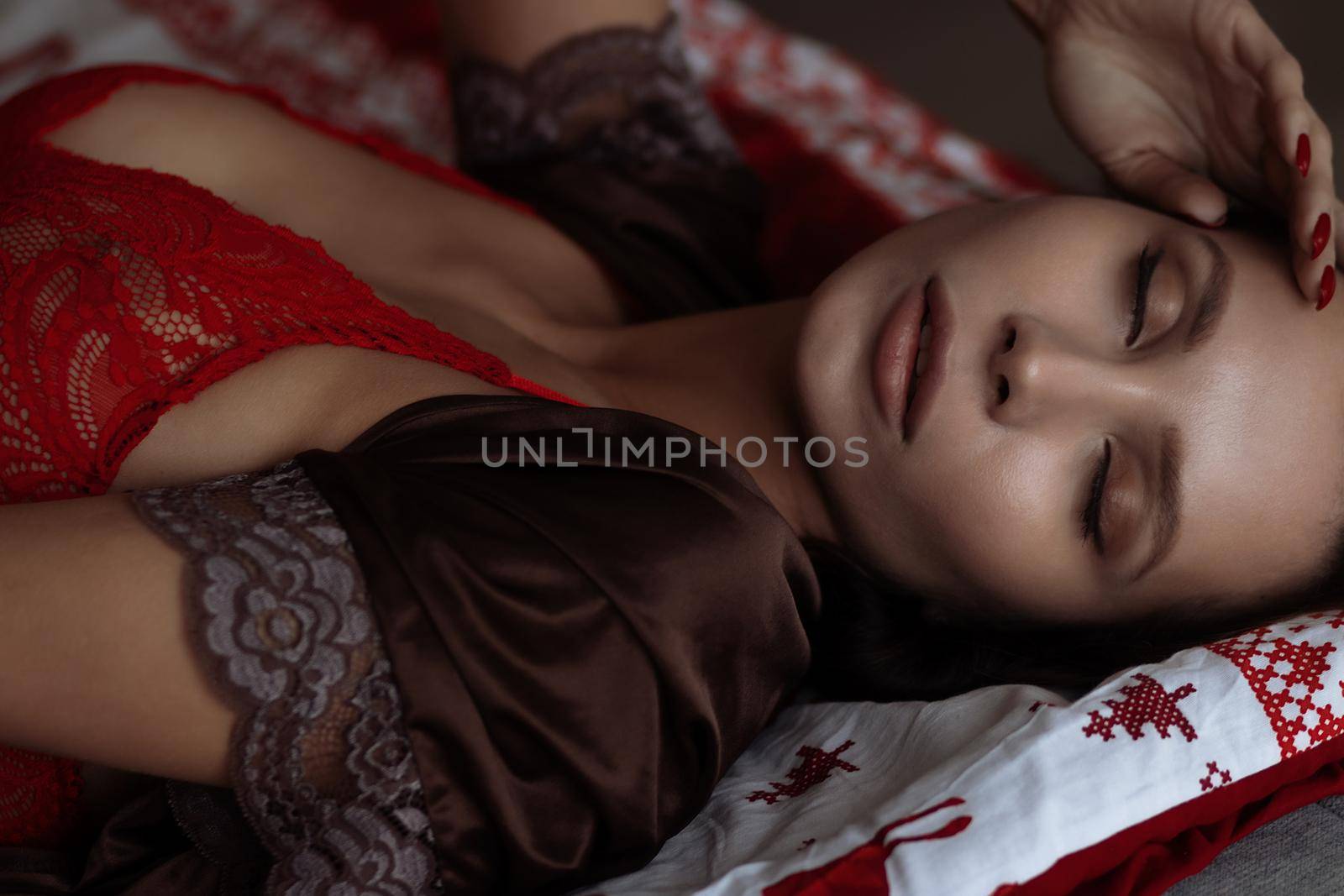 Side view of sexual young female with long dark hair in elegant red bra lying on comfortable bed with closed eyes near decorated Christmas tree