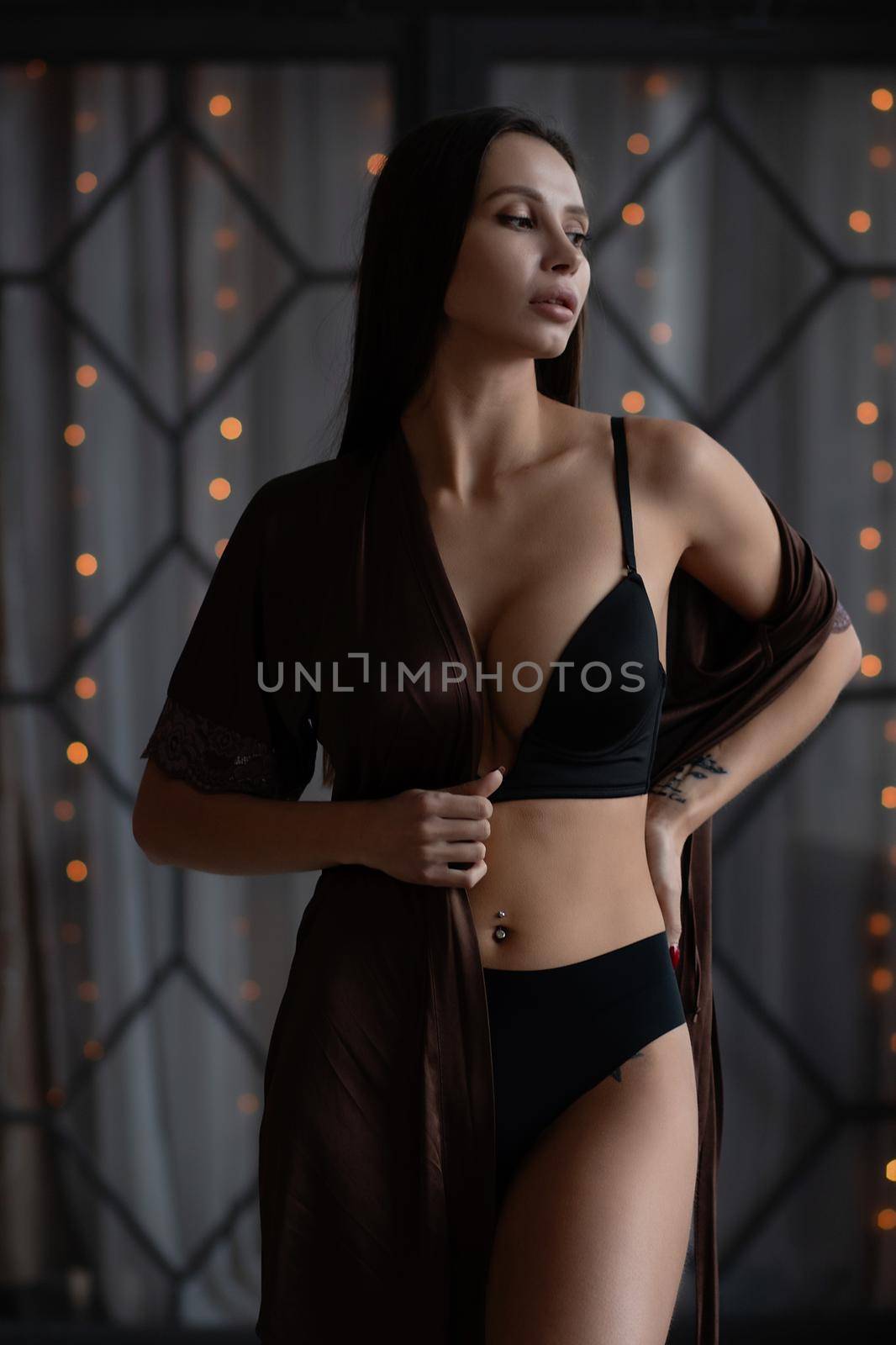 Sensual curvy female in black lingerie and brown silk robe standing gracefully in dark room and looking at camera