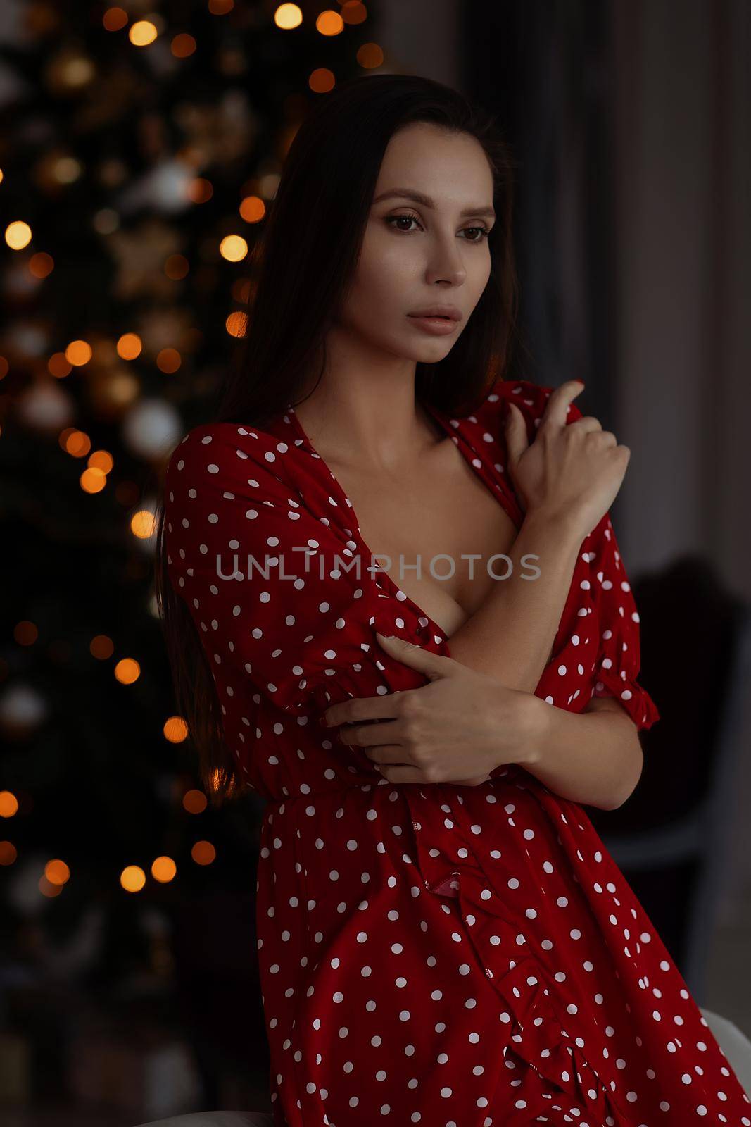 Self esteem young female with long hair in stylish mini dress with decollete standing in dark bedroom with hand on waist and looking at camera