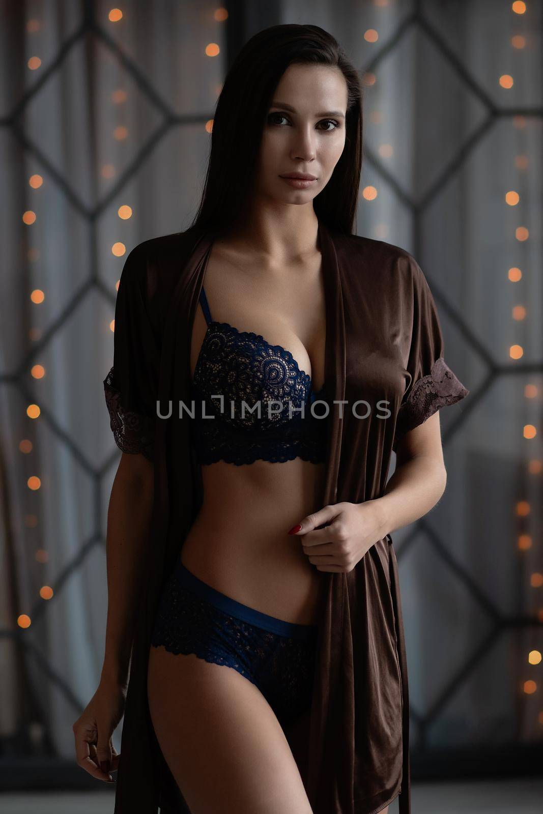 Sensual curvy female in blue lingerie and brown silk robe standing gracefully in dark room and looking at camera