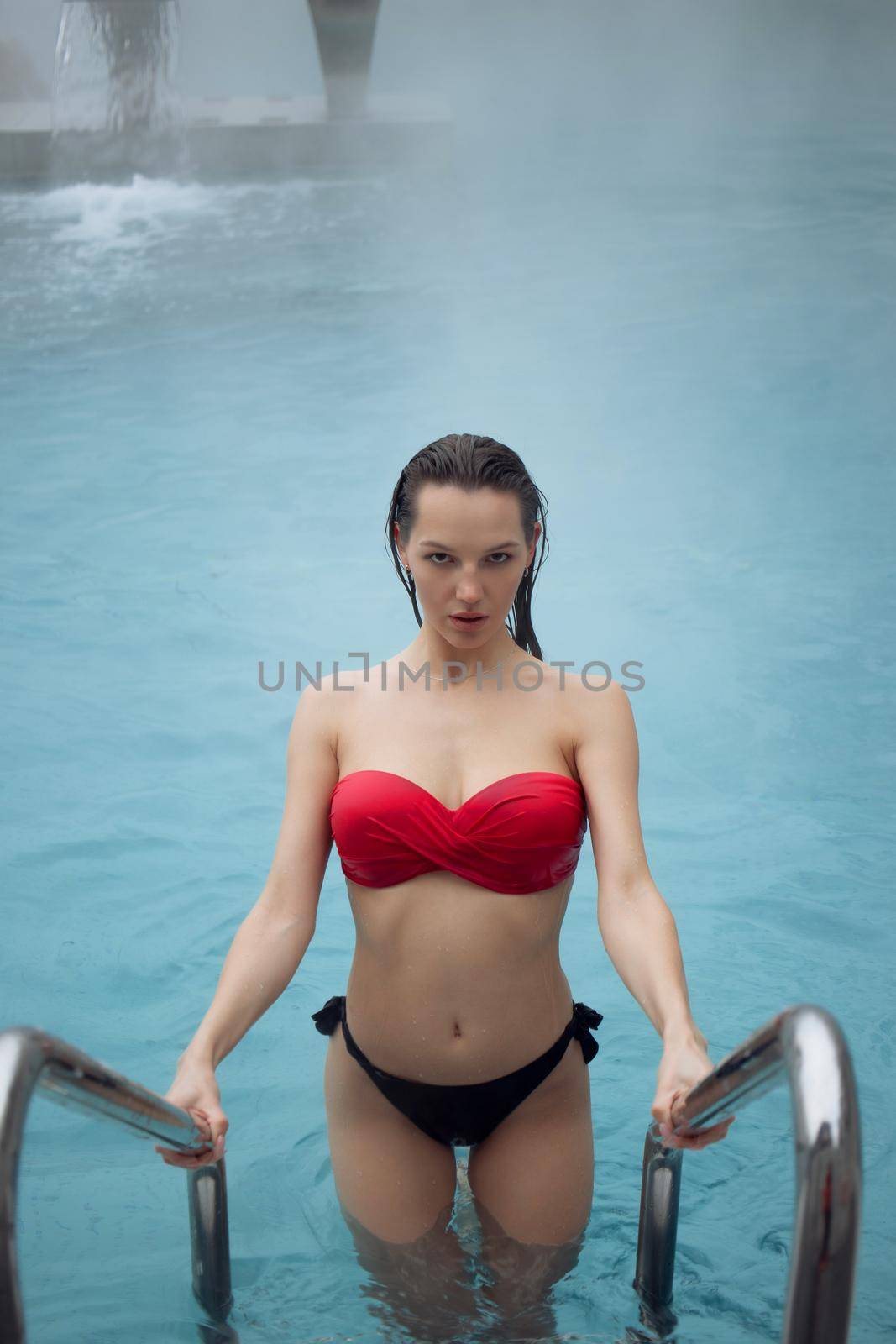 High angle of attractive female in red bikini standing on metal ladder and looking at camera in clean water of pool