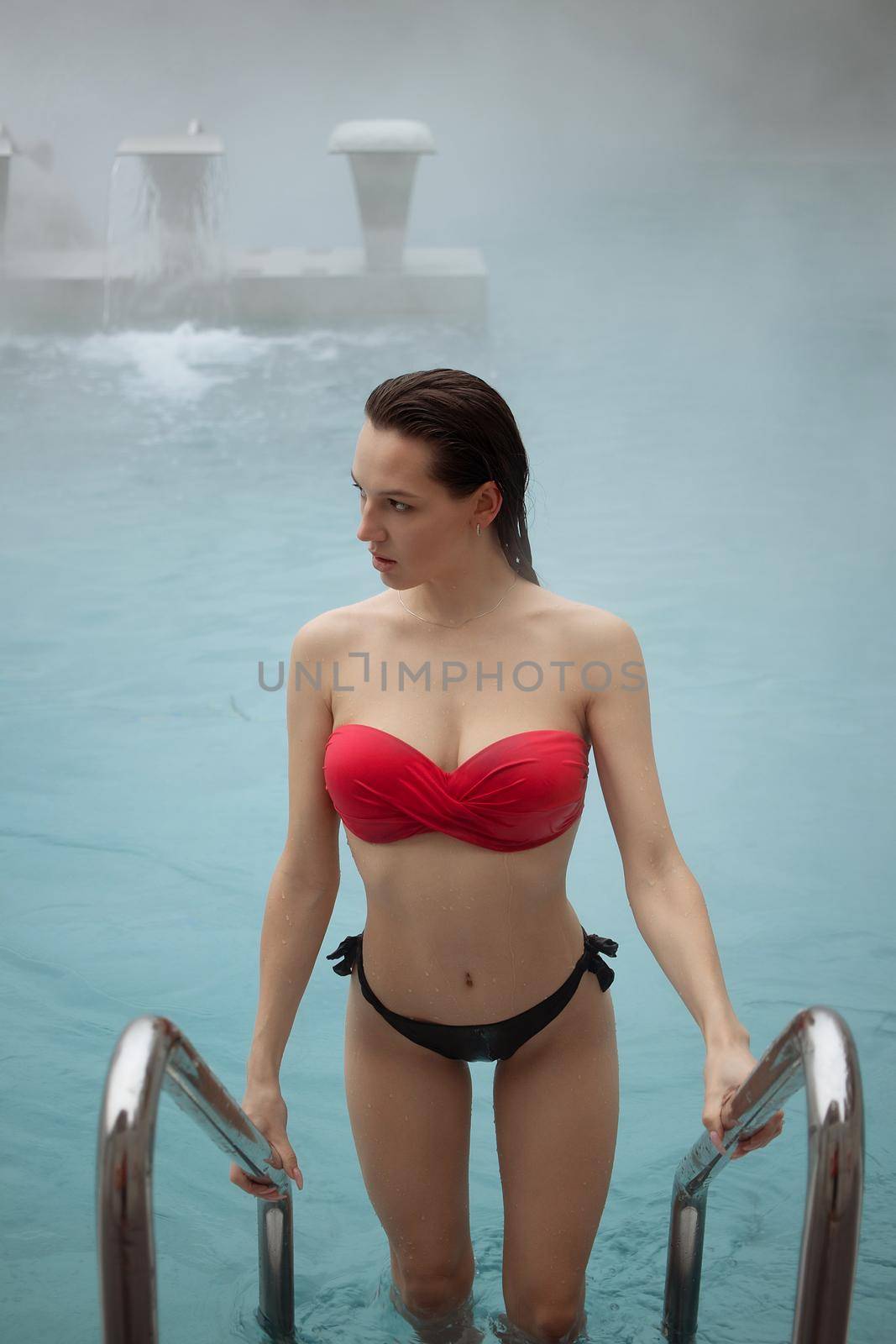 High angle of attractive female in red bikini standing on metal ladder and looking at camera in clean water of pool