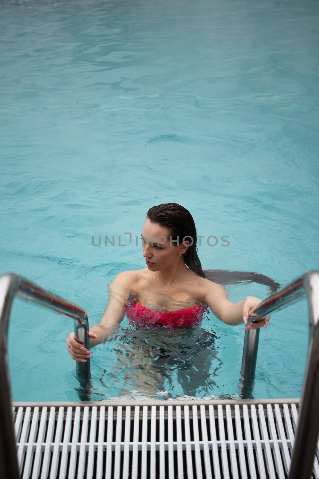 High angle of attractive female in red bikini standing on metal ladder and looking at camera in clean water of pool