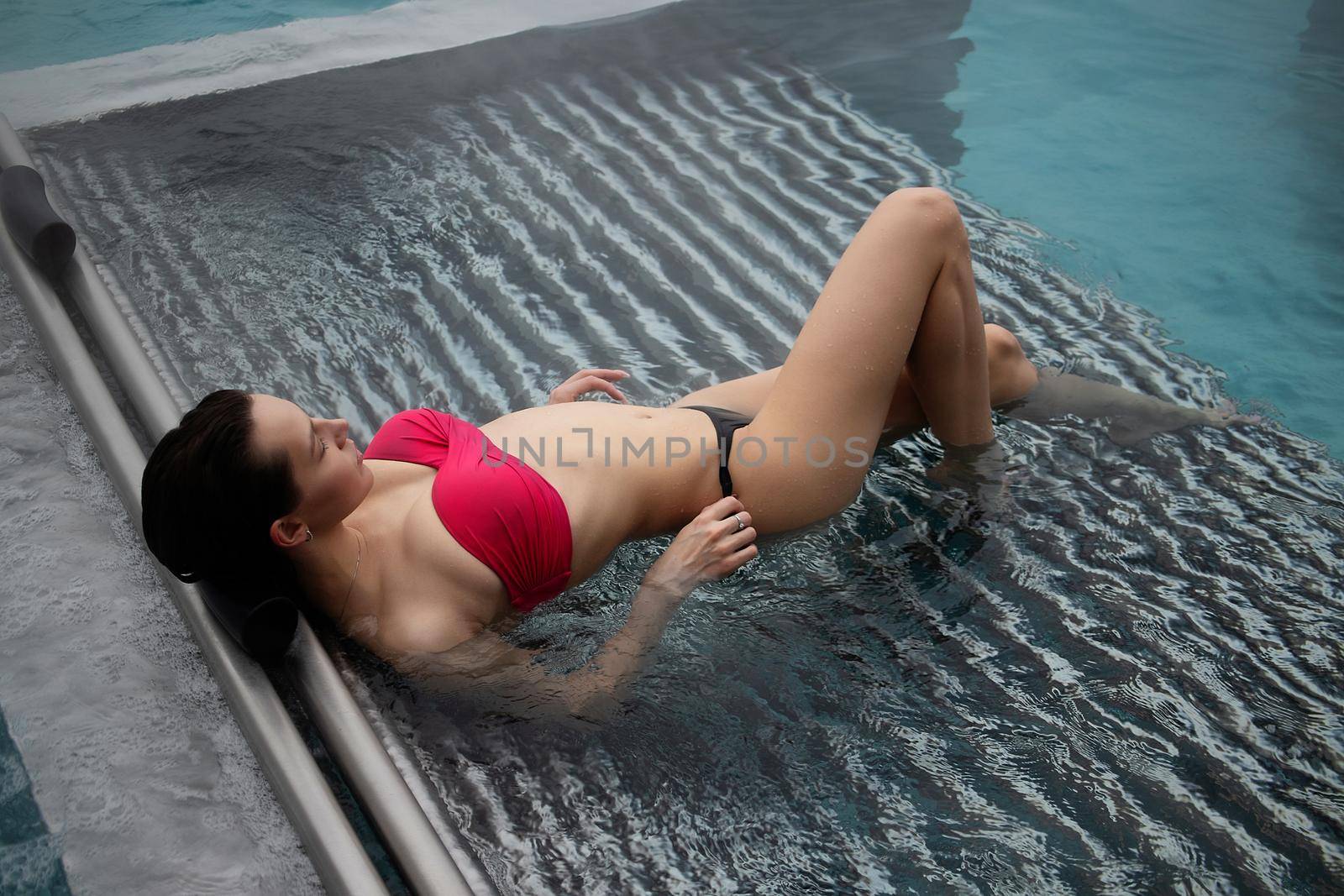 High angle of slim barefoot woman in red swimwear lying on grates in clean water of pool on resort