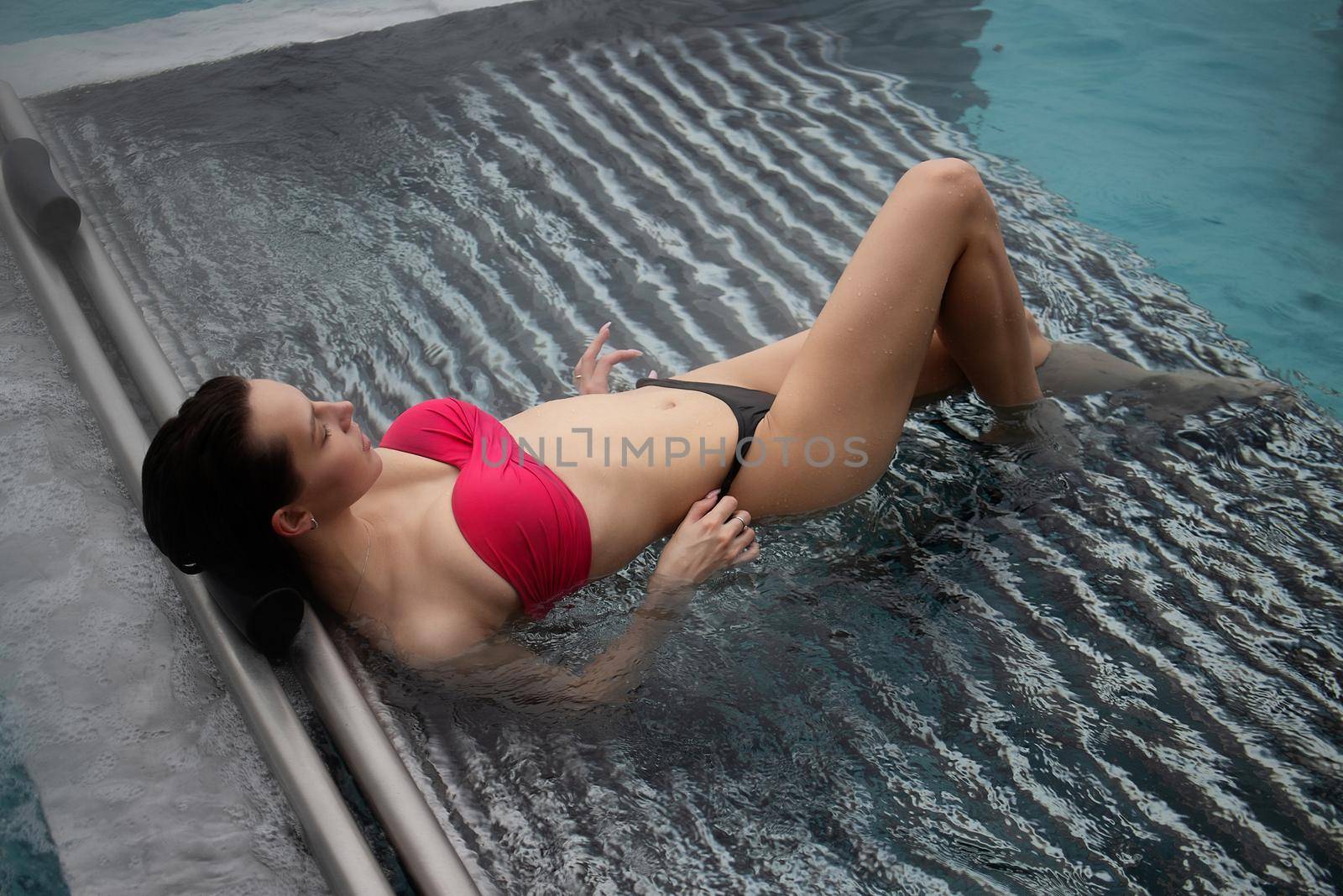 High angle of slim barefoot woman in red swimwear lying on grates in clean water of pool on resort