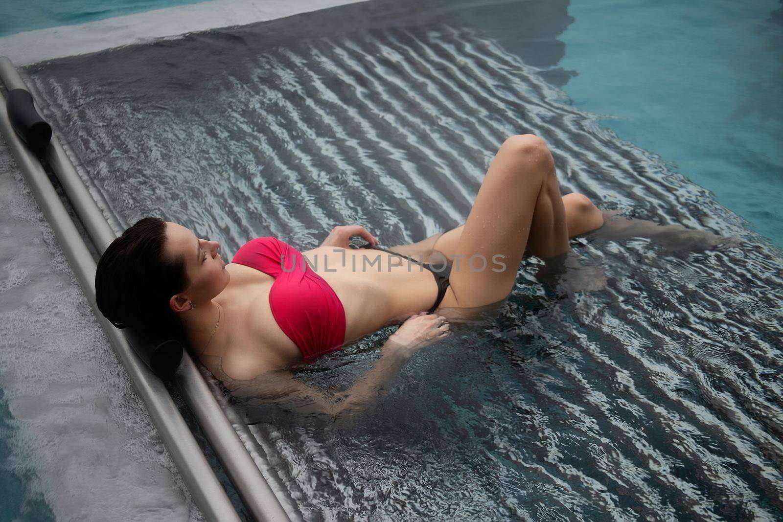 High angle of slim barefoot woman in red swimwear lying on grates in clean water of pool on resort