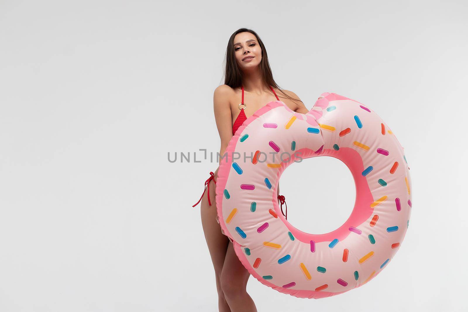 Attractive young slim female in red bikini holding donut colored inflated inflatable ring while standing against white background and representing summer and beach concept