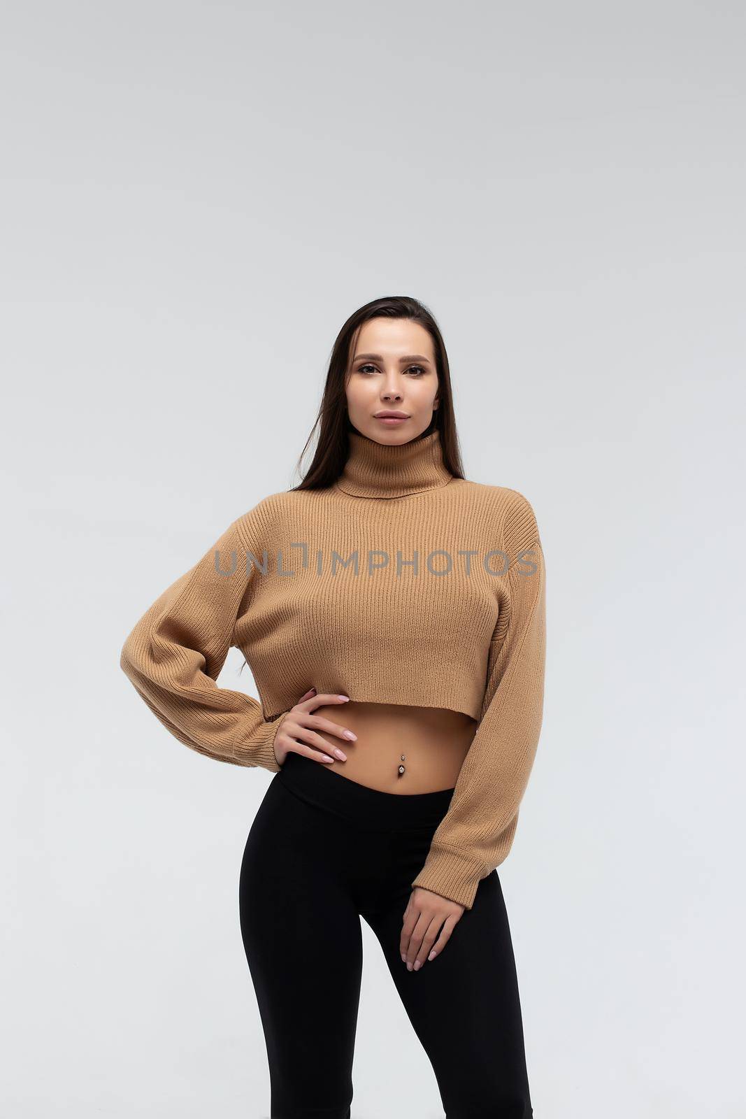 Attractive slim lady in beige jacket and leggings embracing body and looking at camera against white background