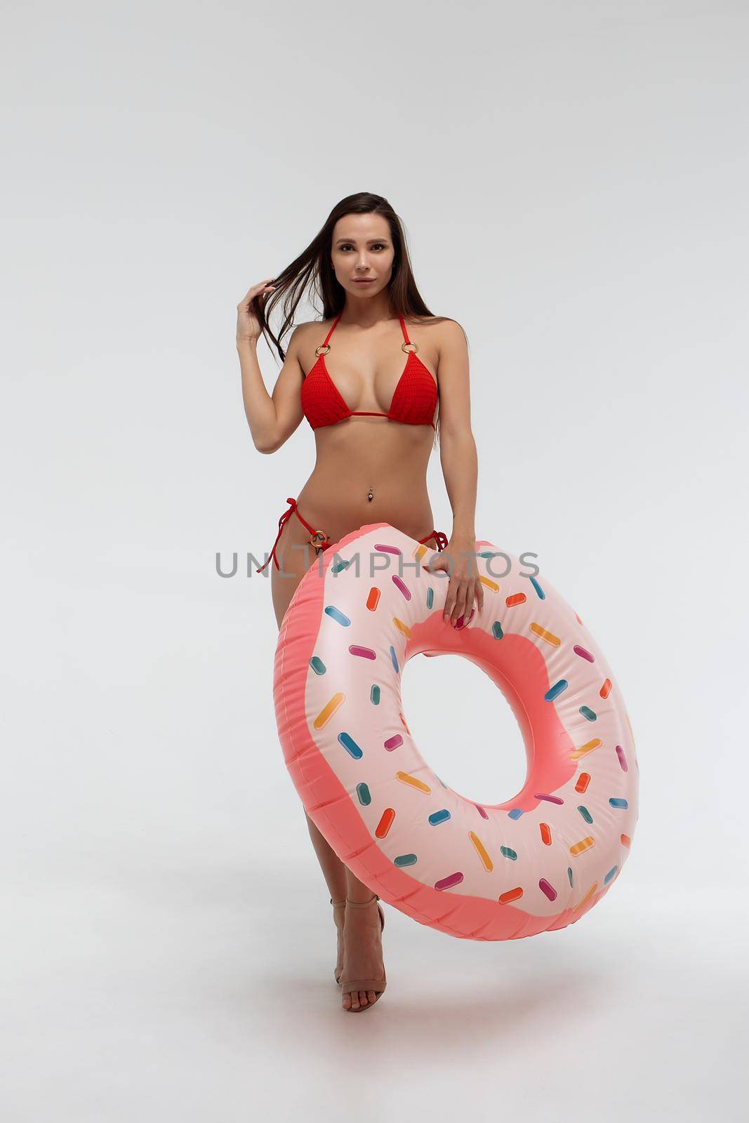 Full body of young slim female model in sexy red bikini carrying donut colored swimming ring and touching hair against white background