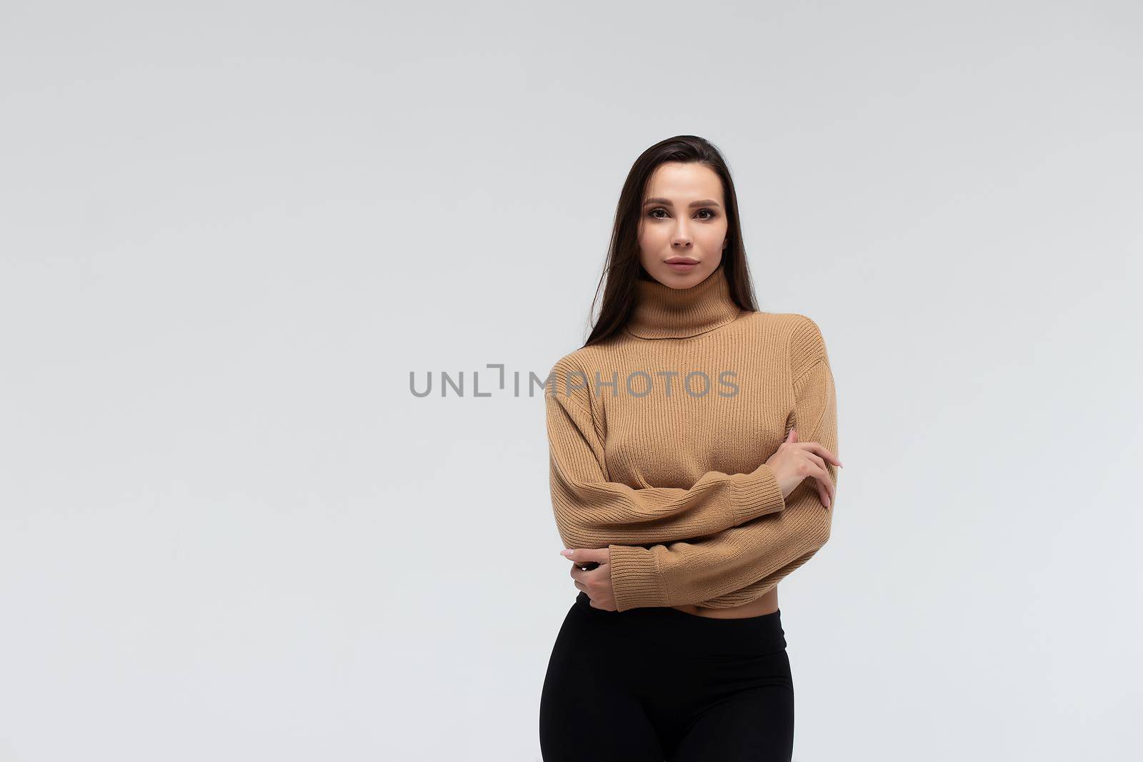 Attractive slim lady in beige jacket and leggings embracing body and looking at camera against white background