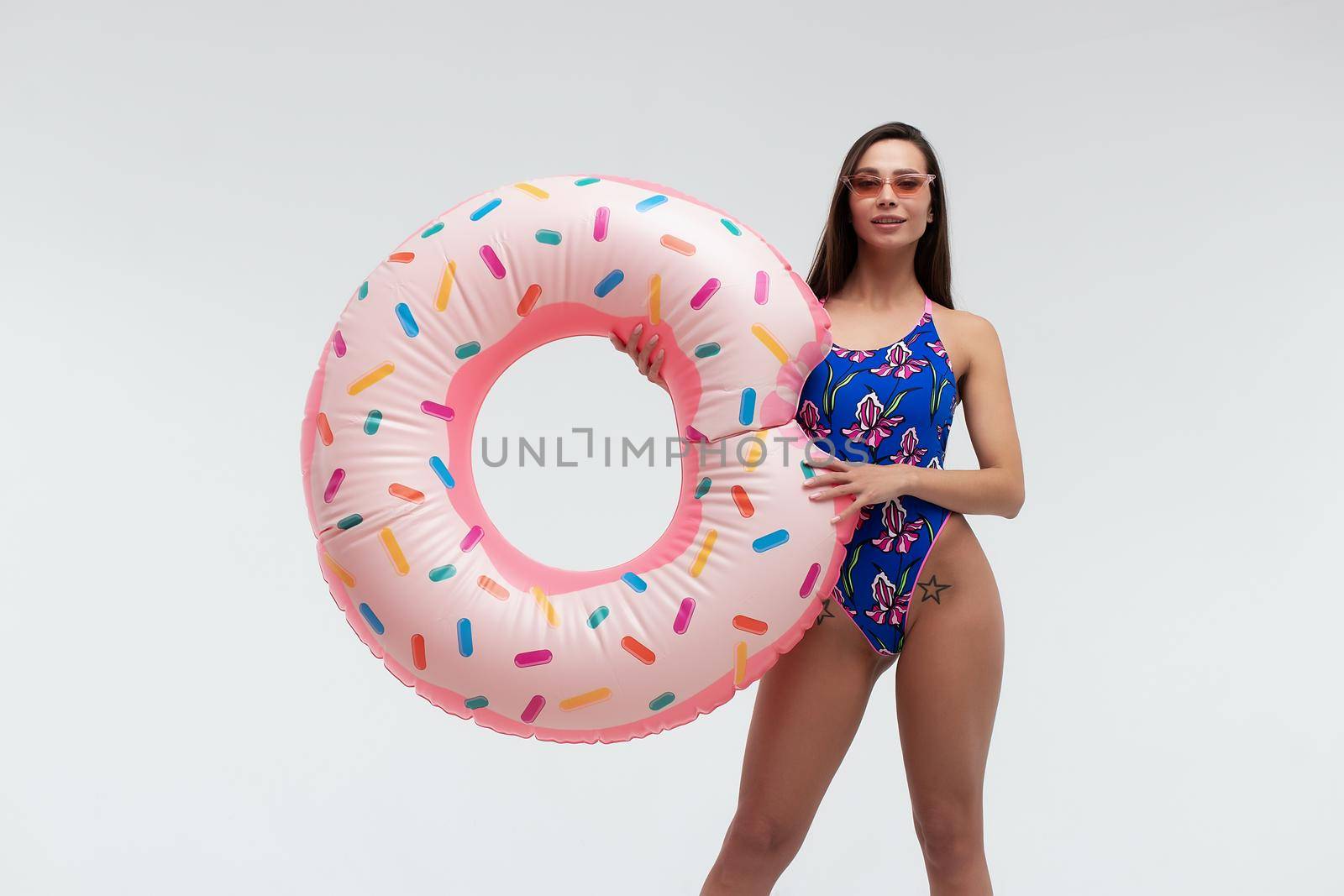 Full body of sexy young long haired brunette in sunglasses wearing stylish swimsuit and high heels holding colorful inflatable ring while standing against white background