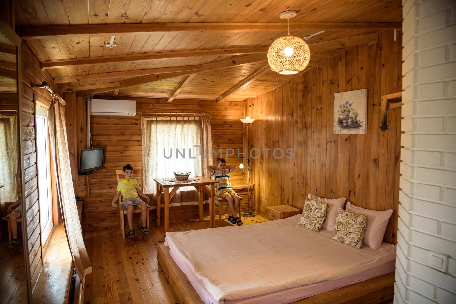 Two boys children in wooden living room in house with contemporary interior design, comfortable bad, carpet on floor, lamplight lamp, decor on table and wooden panel on copy space background.