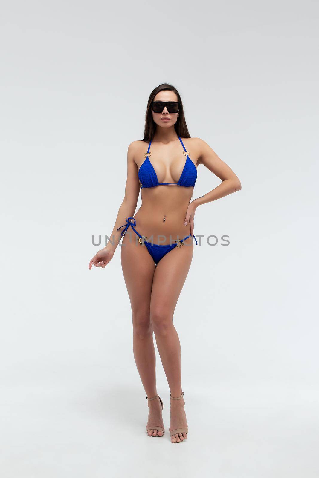 Gorgeous young slim brunette in stylish blue bikini with sunglasses in hand looking at camera on white background