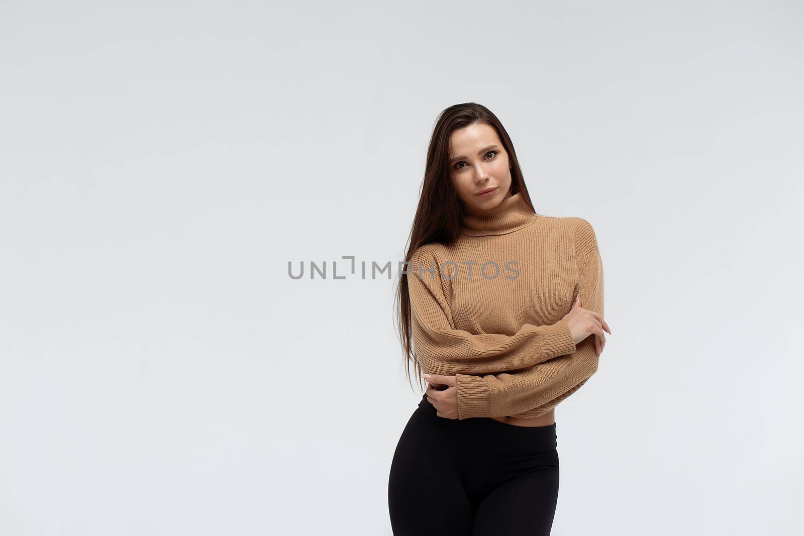 Attractive slim lady in beige jacket and leggings embracing body and looking at camera against white background