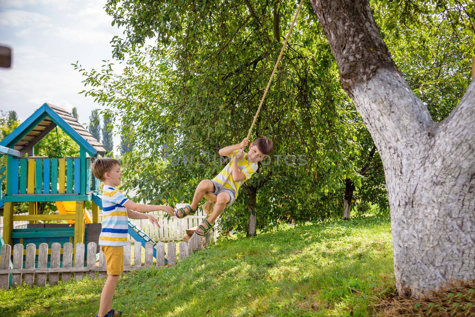 Two adorable happy little boys is having fun on a rope swing which he has found while having rest outside city. Active leisure time with children.