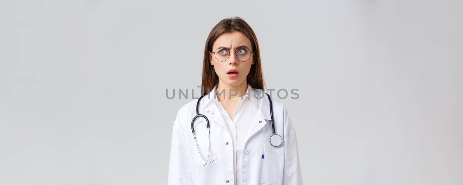 Healthcare workers, medicine, insurance and covid-19 pandemic concept. Shocked and confused young female doctor in white scrubs and glasses, stethoscope, stare left with concerned nervous face.