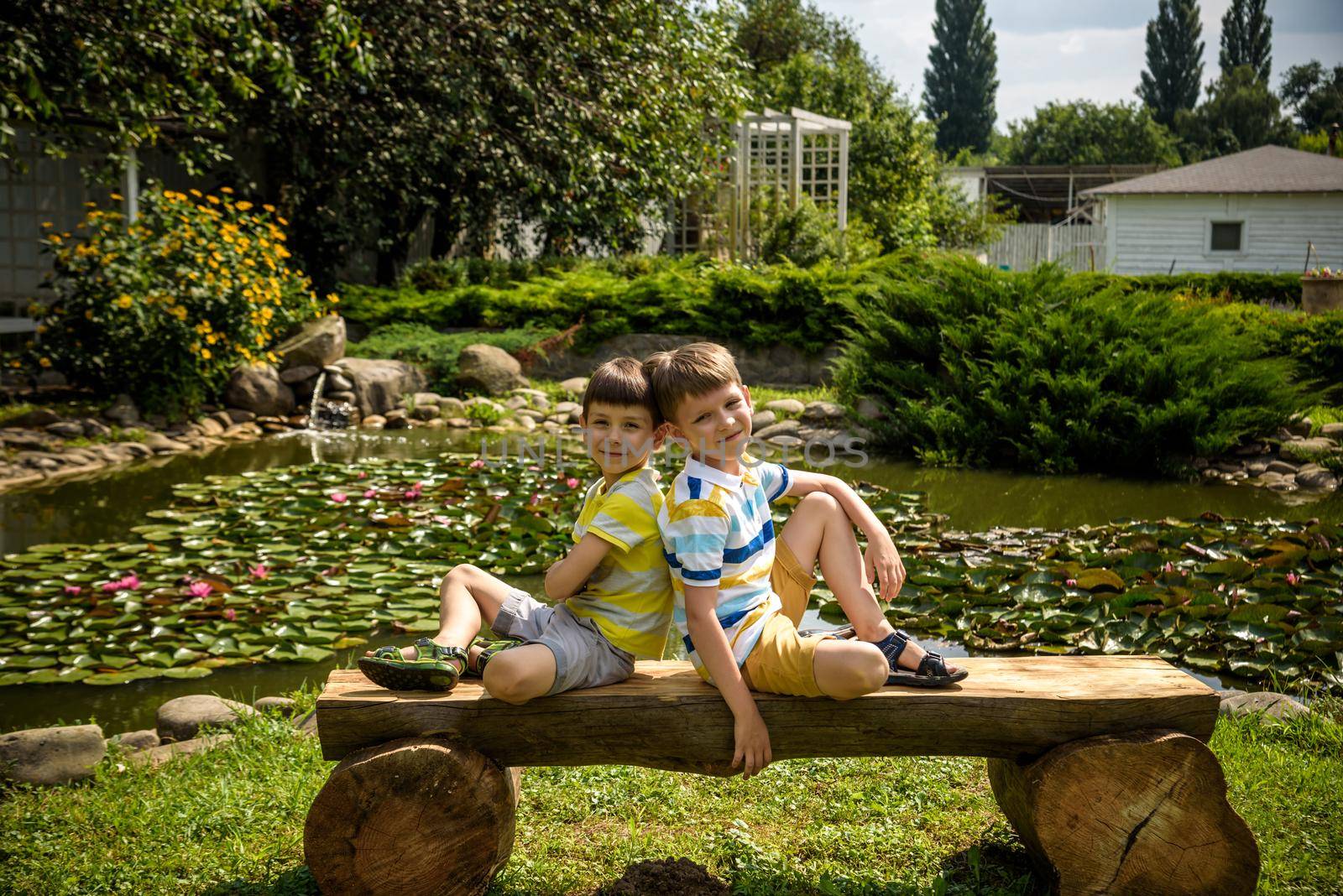 Two brother boys sitting on a bench in a park near peaceful lake with water lily Nymphaeum. Kids relaxing on nature on hot summer day by Kobysh