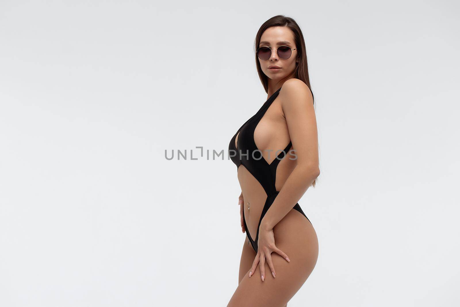 Side view of gorgeous young slim tanned brunette in trendy sunglasses and stylish sexy black swimwear looking at camera