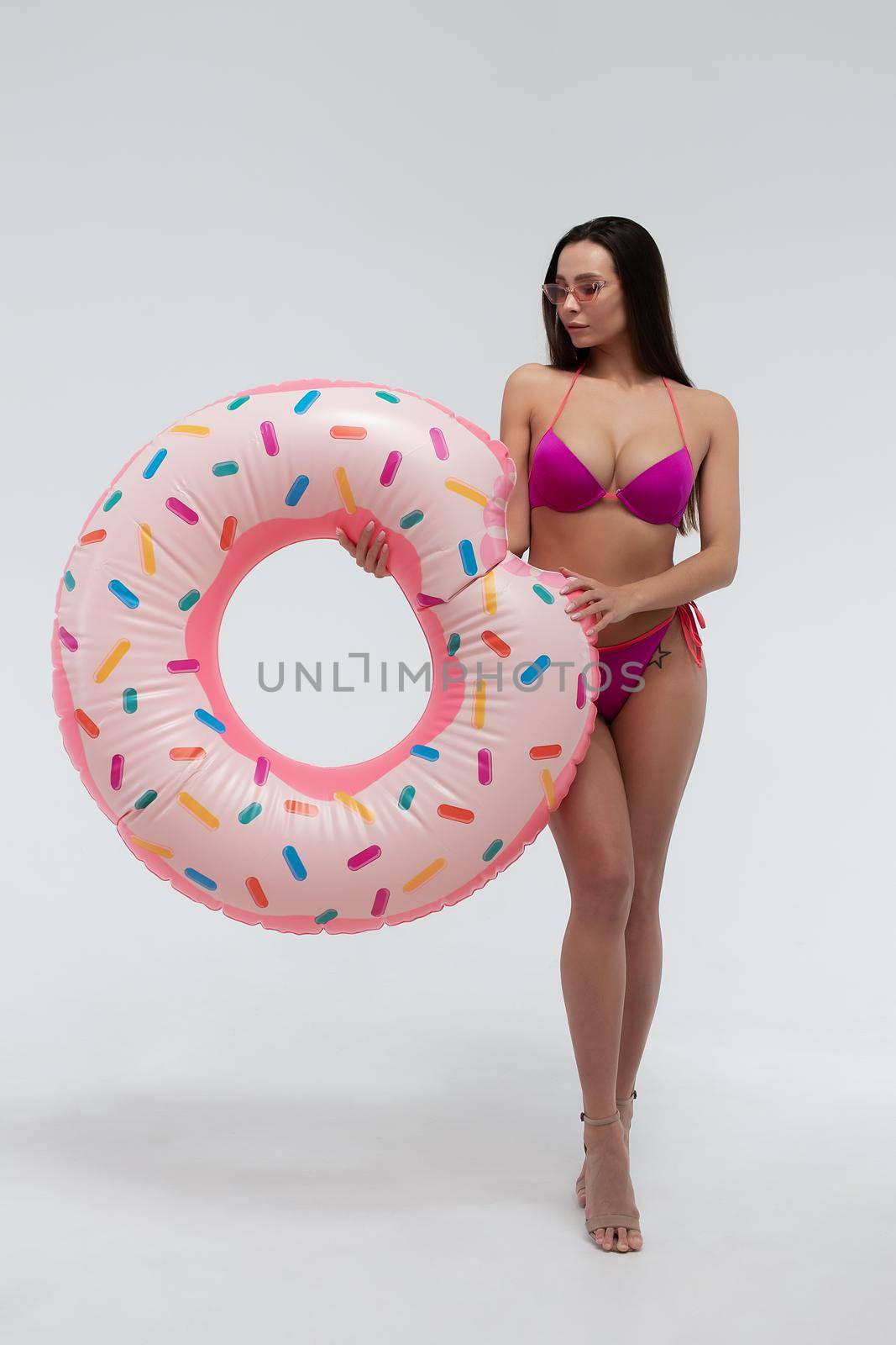 Full body of sexy young long haired brunette in sunglasses wearing stylish swimsuit and high heels holding colorful inflatable ring while standing against white background