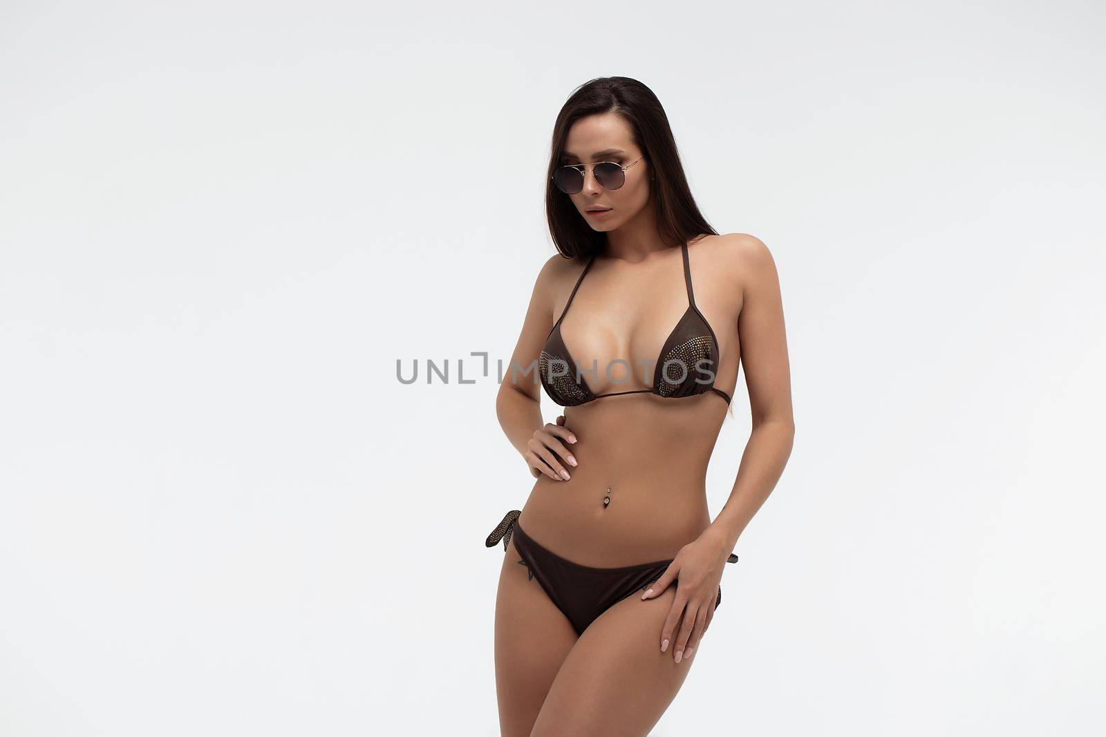 Attractive long haired young brunette model in sexy black bikini and trendy sunglasses touching hair while standing against white background
